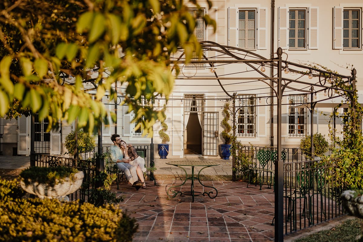 Fotografo matrimonio Rivalba. Villa Ala. Prewedding di Giorgia e Marco. Golden Hour. La coppia è seduti su di una panchina di ferro battuto, davanti alla villa sotto un gazebo in ferro battuto. Ritratto . Giorgia appoggia la testa sulla spalla di Marco.
