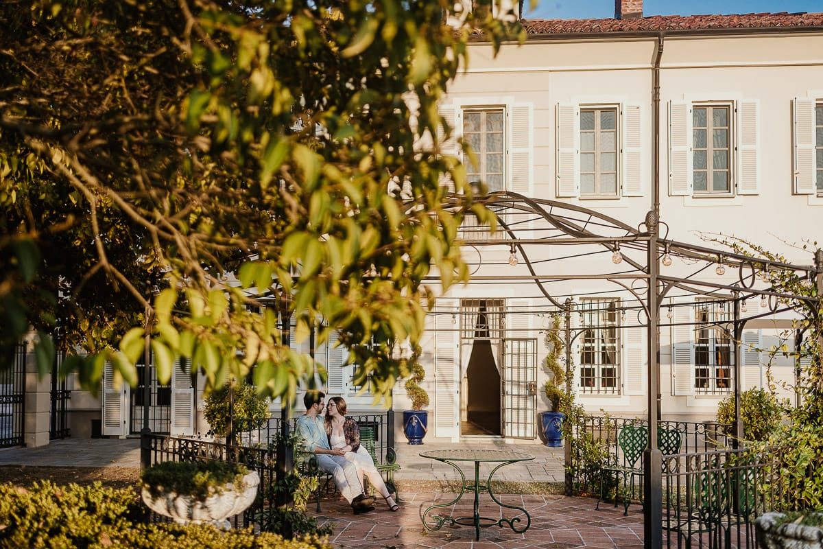 Fotografo matrimonio Rivalba. Villa Ala. Prewedding di Giorgia e Marco. Golden Hour. La coppia è seduti su di una panchina di ferro battuto, davanti alla villa sotto un gazebo in ferro battuto. Ritratto . Giorgia appoggia la testa sulla spalla di Marco.
