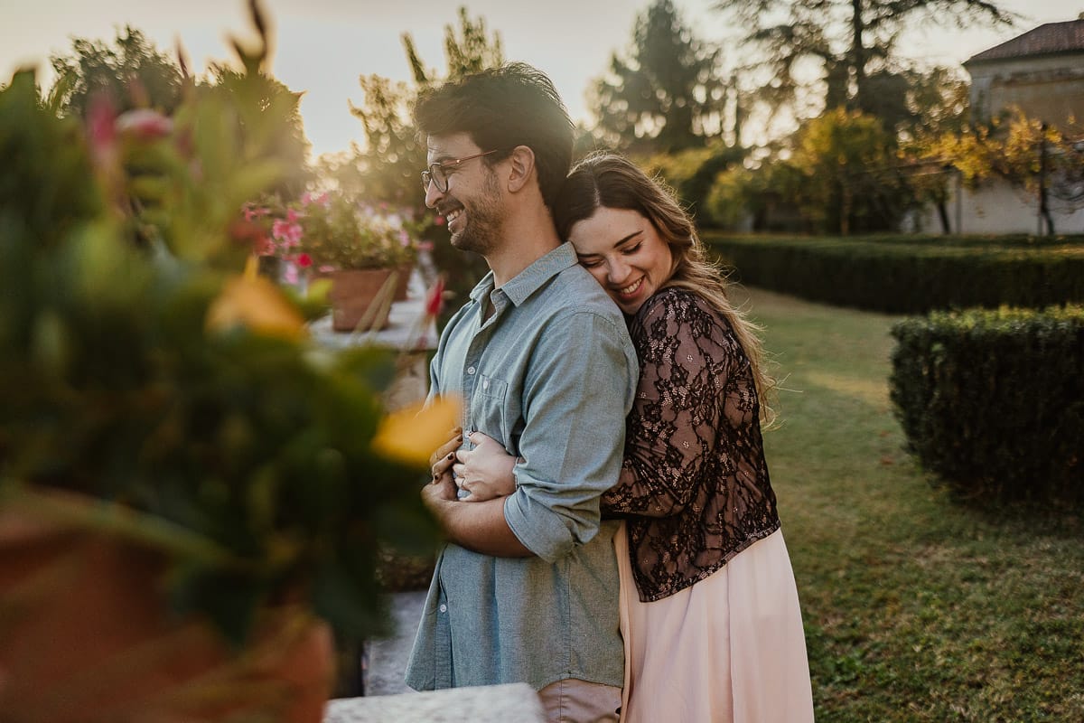 Fotografo matrimonio Rivalba. Villa Ala. Prewedding di Giorgia e Marco. Golden Hour. La coppia è seduti su di una panchina di ferro battuto, davanti alla villa sotto un gazebo in ferro battuto. Ritratto. Giorgia è dietro Marco e si appoggia teneramente a lui.