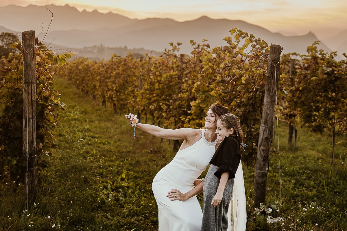 Fotografo matrimonio Torino. Matrimonio eco-consapevole a Cascina Ranverso. Ritratto degli sposi al tramonto. Sposi nel vigneto la sposa si fa un selfie con la cuginetta