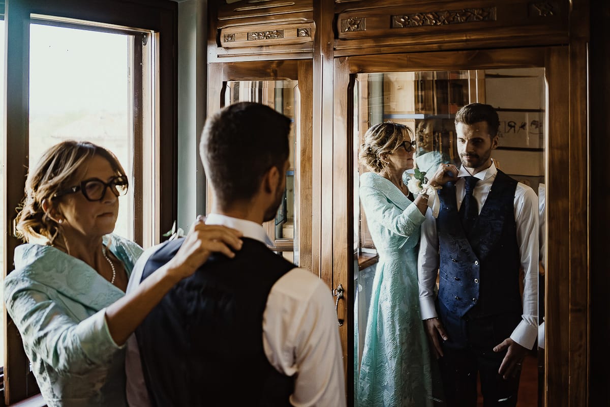 Fotografo matrimonio Torino. Matrimonio eco-consapevole a Cascina Ranverso. Preparazione dello sposo a Chieri. Vestizione dello sposo. La mamma aiuta lo sposo riflessi nelle ante a vetro dell'armadio.