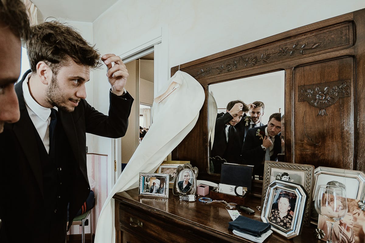 Fotografo matrimonio Torino. Matrimonio eco-consapevole a Cascina Ranverso. Preparazione dello sposo a Chieri. Vestizione dello sposo. Sposo con gli amici riflessi in uno specchio.