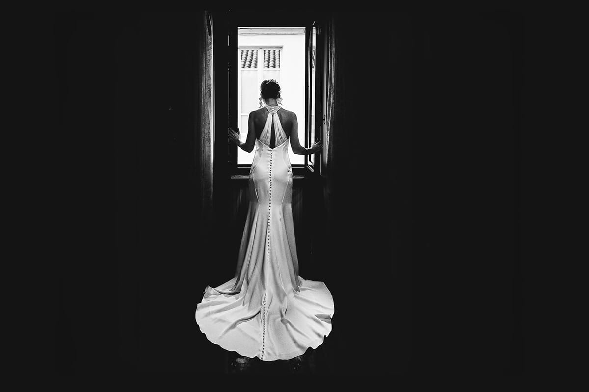 Fotografo matrimonio Torino. Matrimonio eco-consapevole a Cascina Ranverso. Preparazione della sposa a Chieri. Sposa alla finestra di schiena. Bianco e nero
