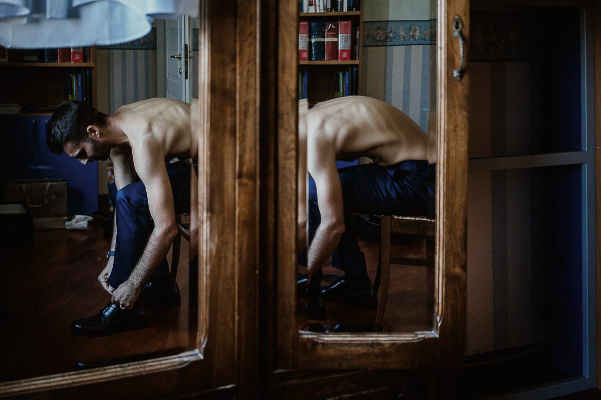 Fotografo matrimonio Torino. Matrimonio eco-consapevole a Cascina Ranverso. Preparazione dello sposo a Chieri. Vestizione dello sposo. Lo sposo si mette le scarpe riflesso nelle ante a vetro di un armadio