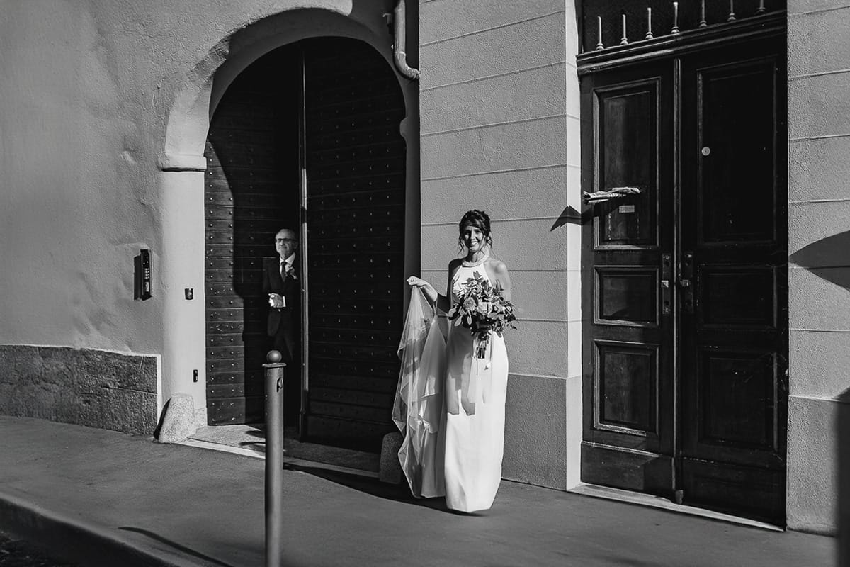 Fotografo matrimonio Torino. Matrimonio eco-consapevole a Cascina Ranverso. Preparazione della sposa a Chieri. Sposa fuori dal portene di casa attende l'auto. Il papà fa capolino dal portone. Bianco e nero.