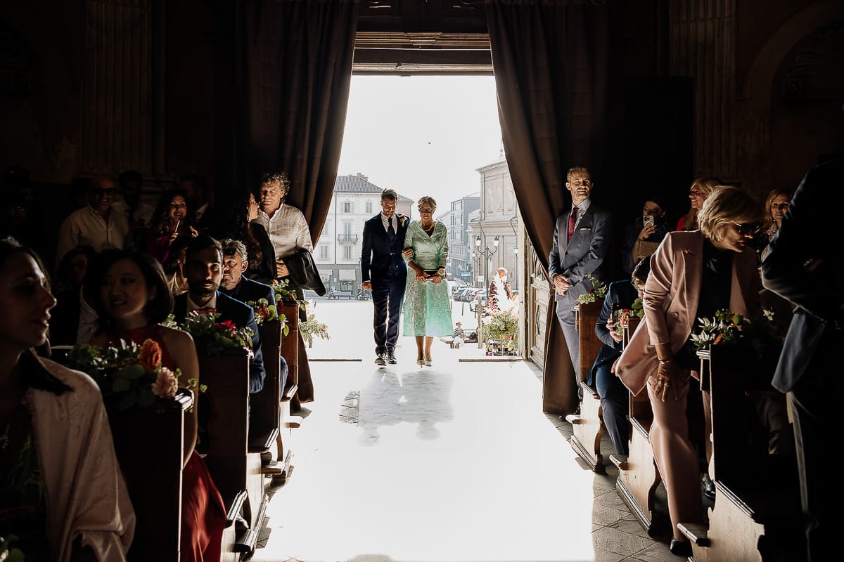 Fotografo matrimonio Torino. Matrimonio eco-consapevole a Cascina Ranverso. Cerimonia a Chieri nella chiesa dei Santi Bernardino e Rocco. Sposo sta per entrare con la madre.