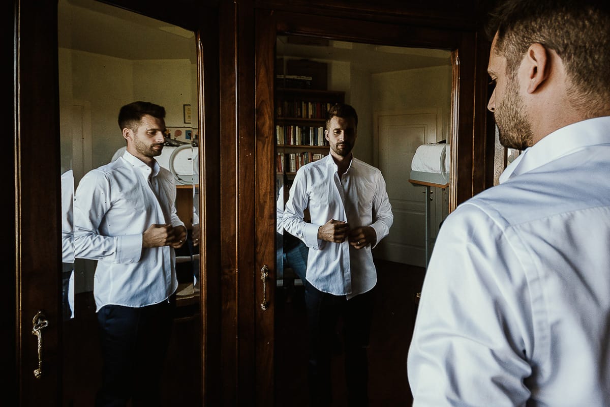 Fotografo matrimonio Torino. Matrimonio eco-consapevole a Cascina Ranverso. Preparazione dello sposo a Chieri. Vestizione dello sposo. Lo sposo indossa la camicia riflesso nelle ante a vetro dell'armadio