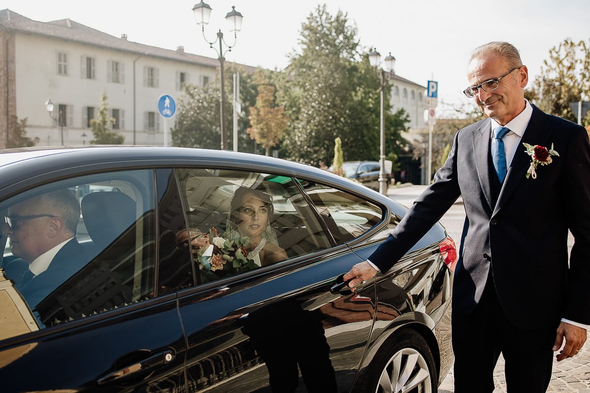 Fotografo matrimonio Torino. Matrimonio eco-consapevole a Cascina Ranverso. Cerimonia a Chieri nella chiesa dei Santi Bernardino e Rocco. Arrivo della sposa. Il padre, riflesso nell'auto si appresta ad aprire la portiera per far scendere la sposa.