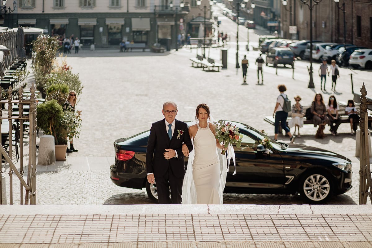 Fotografo matrimonio Torino. Matrimonio eco-consapevole a Cascina Ranverso. Cerimonia a Chieri nella chiesa dei Santi Bernardino e Rocco. Sposa e padre fuori dall'auto. Salgono le scale della chiesa.