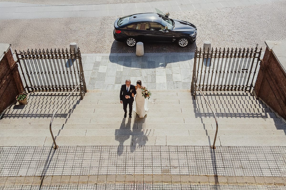 Fotografo matrimonio Torino. Matrimonio eco-consapevole a Cascina Ranverso. Cerimonia a Chieri nella chiesa dei Santi Bernardino e Rocco. Sposa e padre fuori dall'auto. Salgono le scale della chiesa. Foto dal drone