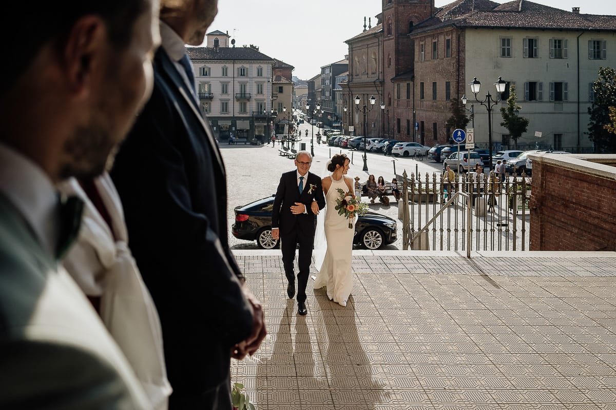 Fotografo matrimonio Torino. Matrimonio eco-consapevole a Cascina Ranverso. Cerimonia a Chieri nella chiesa dei Santi Bernardino e Rocco. Sposa e padre fuori dall'auto. Salgono le scale della chiesa.