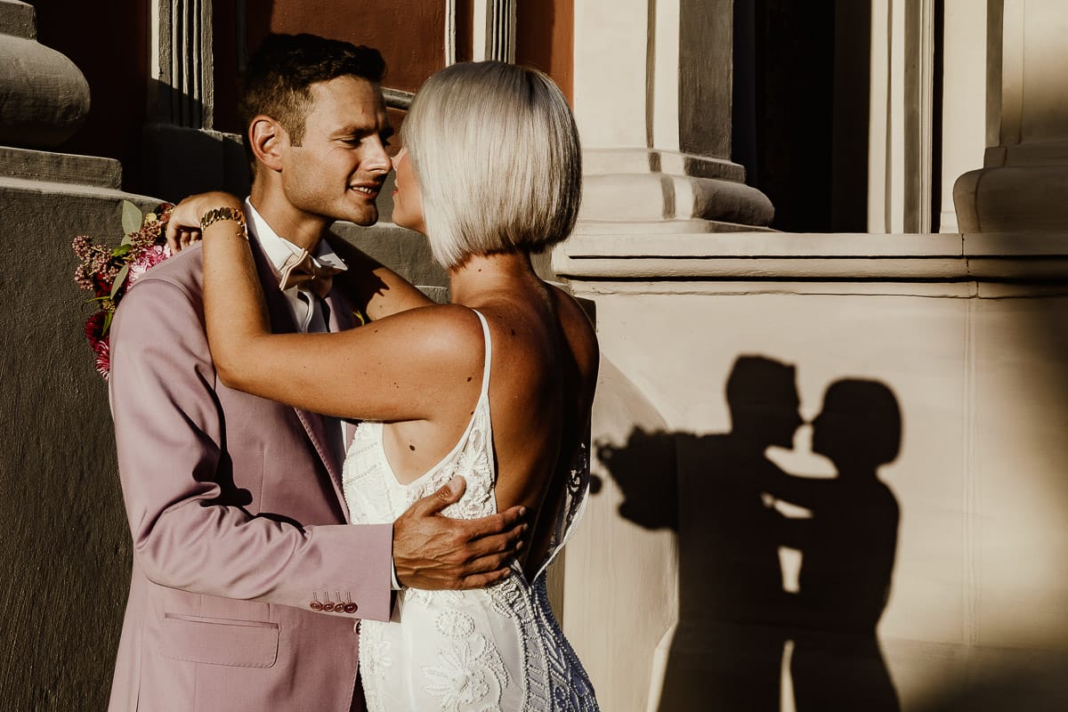 Matrimonio boho-chic da favola nel Monferrato. Fotografo matrimonio Monferrato. matrimonio a Molare (AL). Servizio fotografico di coppia degli sposi. sposi si baciano. Il sole proietta la loro ombra sul muro.