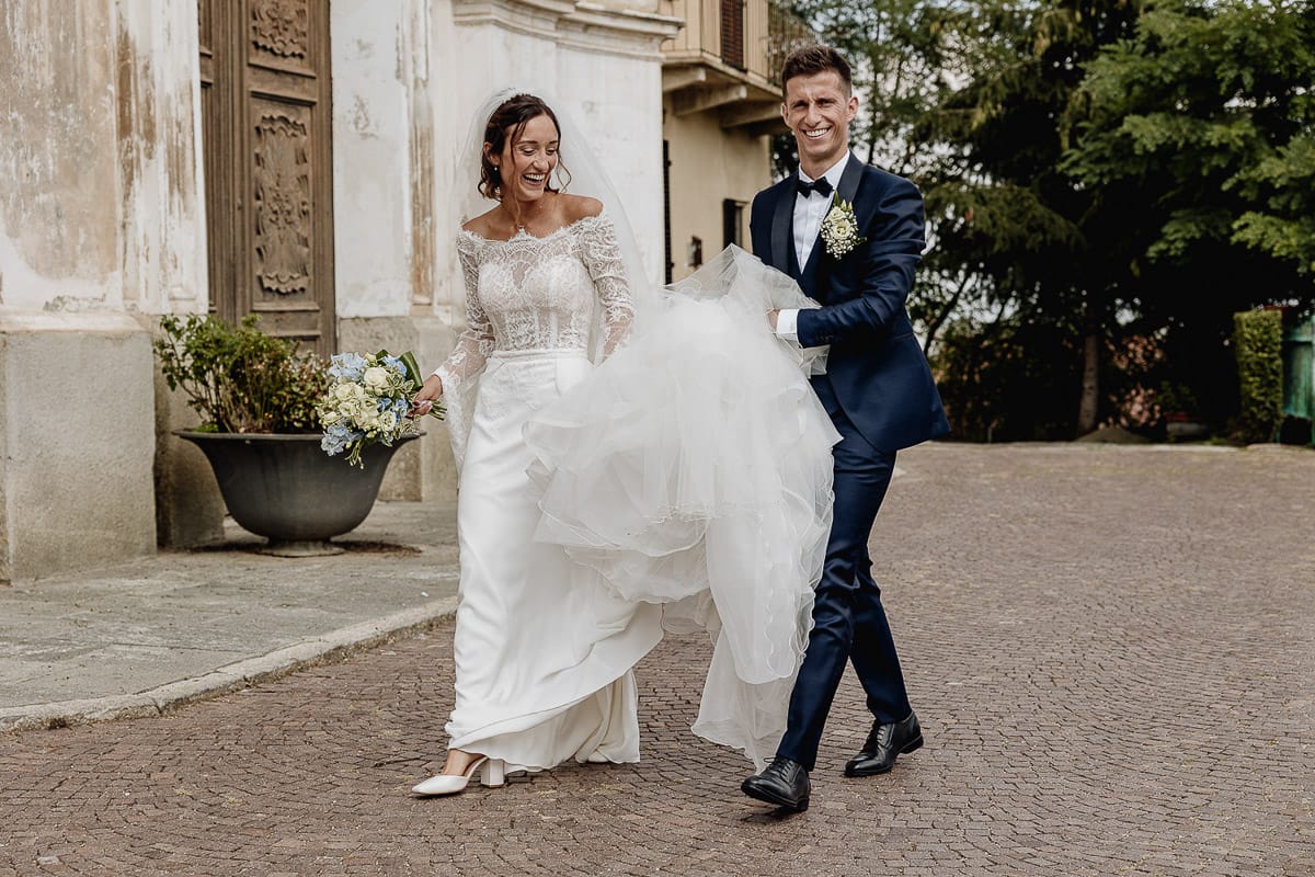 Fotografo matrimonio Torino | Tenuta La Michelina | Pinerolo. Ritratto di coppia degli sposi. Sposi camminano. lo sposo tiene l'abito della sposa. Entrambi ridono.