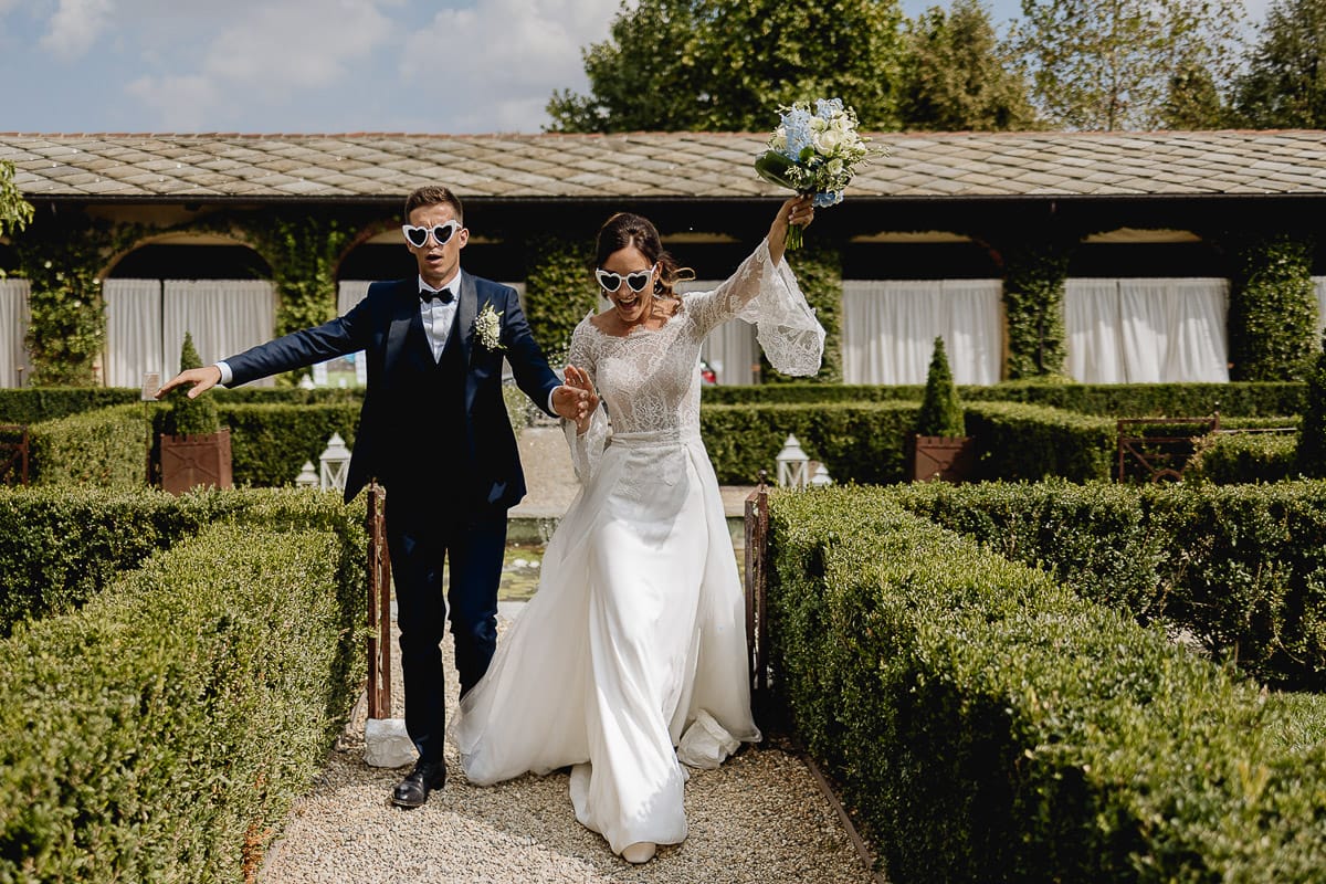 Fotografo matrimonio Torino | Tenuta La Michelina | Pinerolo. Sposi corrono nel vialetto del giardino all'italiana, con gli occhiali da sole a forma di cuore.