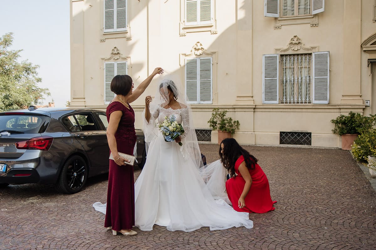 Fotografo matrimonio Torino | Tenuta La Michelina | Pinerolo. Cerimonia religiosa nella chiesa di Santa Maria Maggiore di Poirino. Ritratto della sposa che si dirige verso la chiesa. mamma le sistema il velo e amica le sistema l'abito.