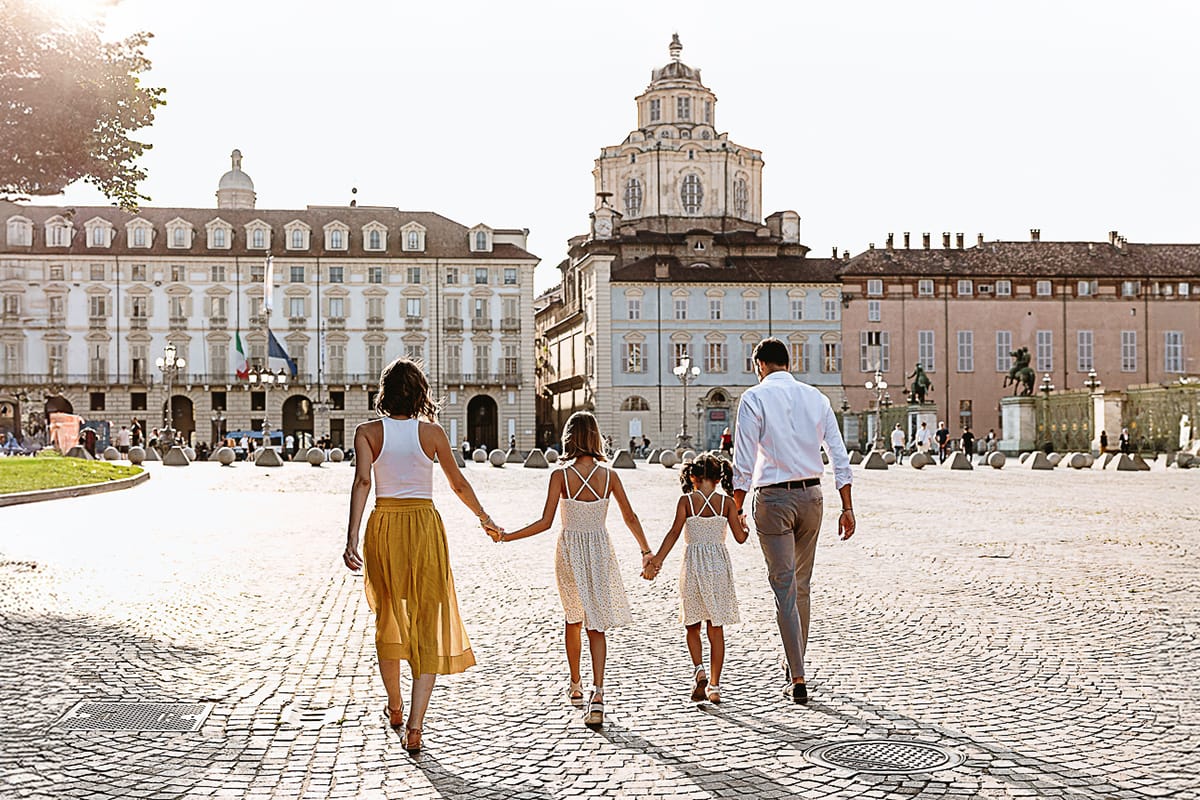 Fotografia di famiglia in vacanza a Torino: famiglia americana vista da dietro passeggia in piazza Castello tenendosi per mano.