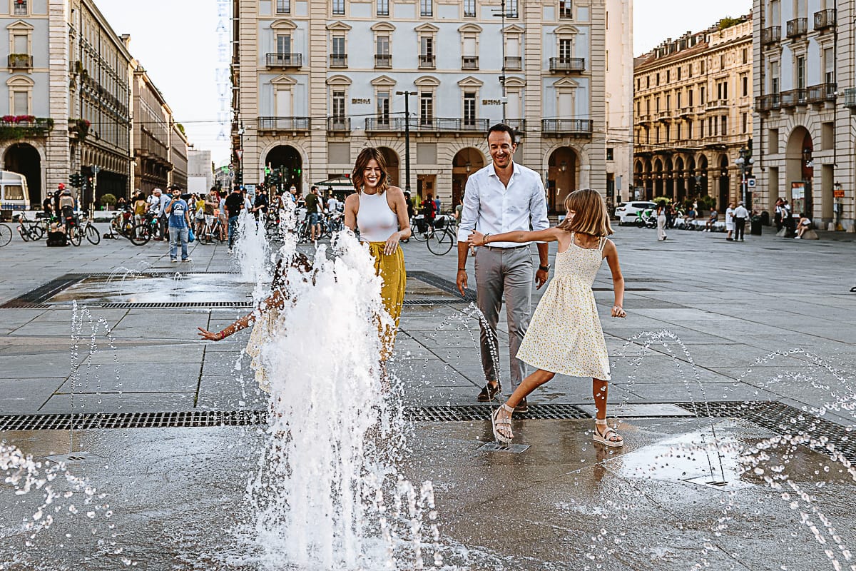 Fotografia di famiglia in vacanza a Torino: bambine giocano con l'acqua delle fontane di piazza Castello, i genitori le guardano