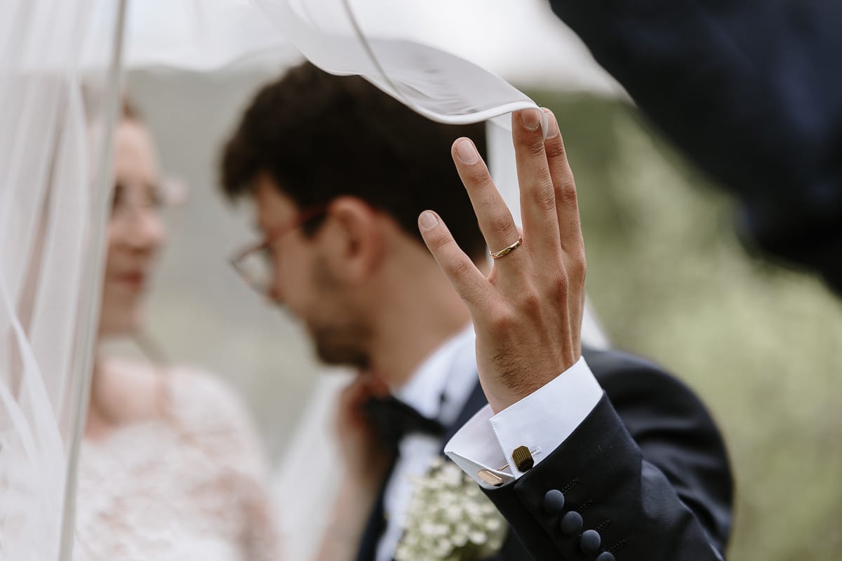 Fotografo matrimonio Torino. Sposi sotto il velo nel giardino di Villa Ala. lo sposo tiene il velo, particolare della fede nuziale