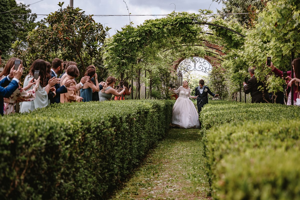 Fotografo matrimonio Torino. Sposi entrano nella zona a peritivo passando sotto un arco con i rampicanti. Gli invitati applaudono e scattano foto con il cellulare.