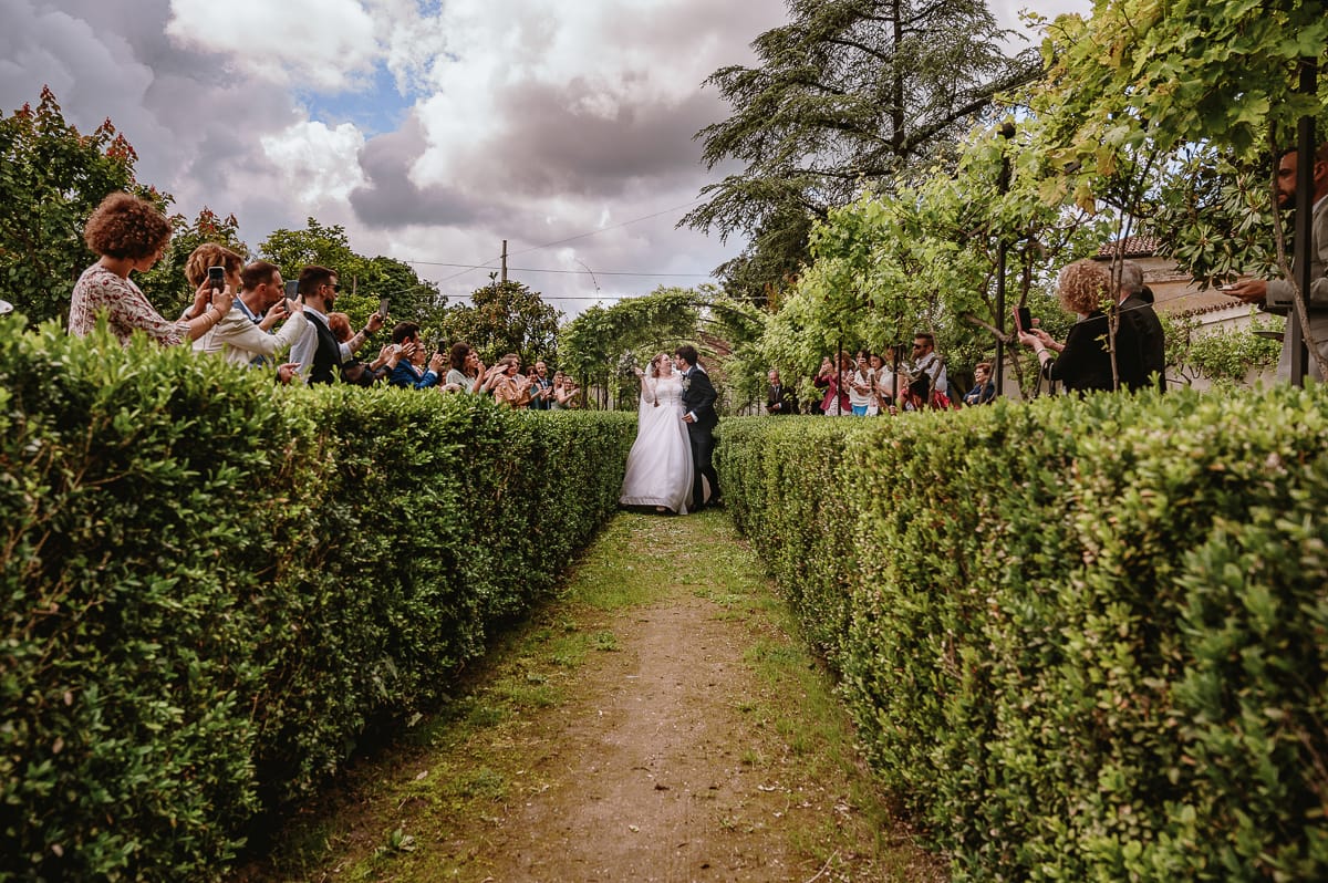 Fotografo matrimonio Torino. Sposi si baciano nella zona aperitivo passando sotto un arco con i rampicanti. Gli invitati applaudono e scattano foto con il cellulare.