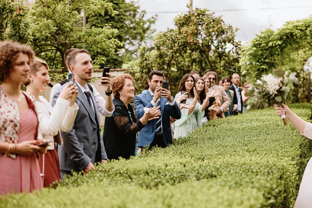 Fotografo matrimonio Torino. Aperitivo. Gli sposi passano sotto un arco con i ram. Gli invitati applaudono e scattano foto con il cellulare.