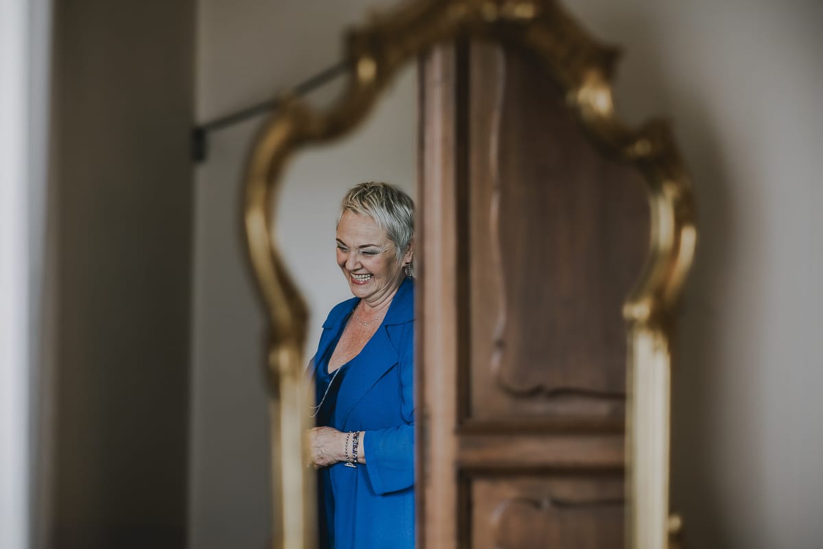 Preparazione della sposa a Madonna della Scala. Mamma della sposa che ride riflessa in uno specchio.