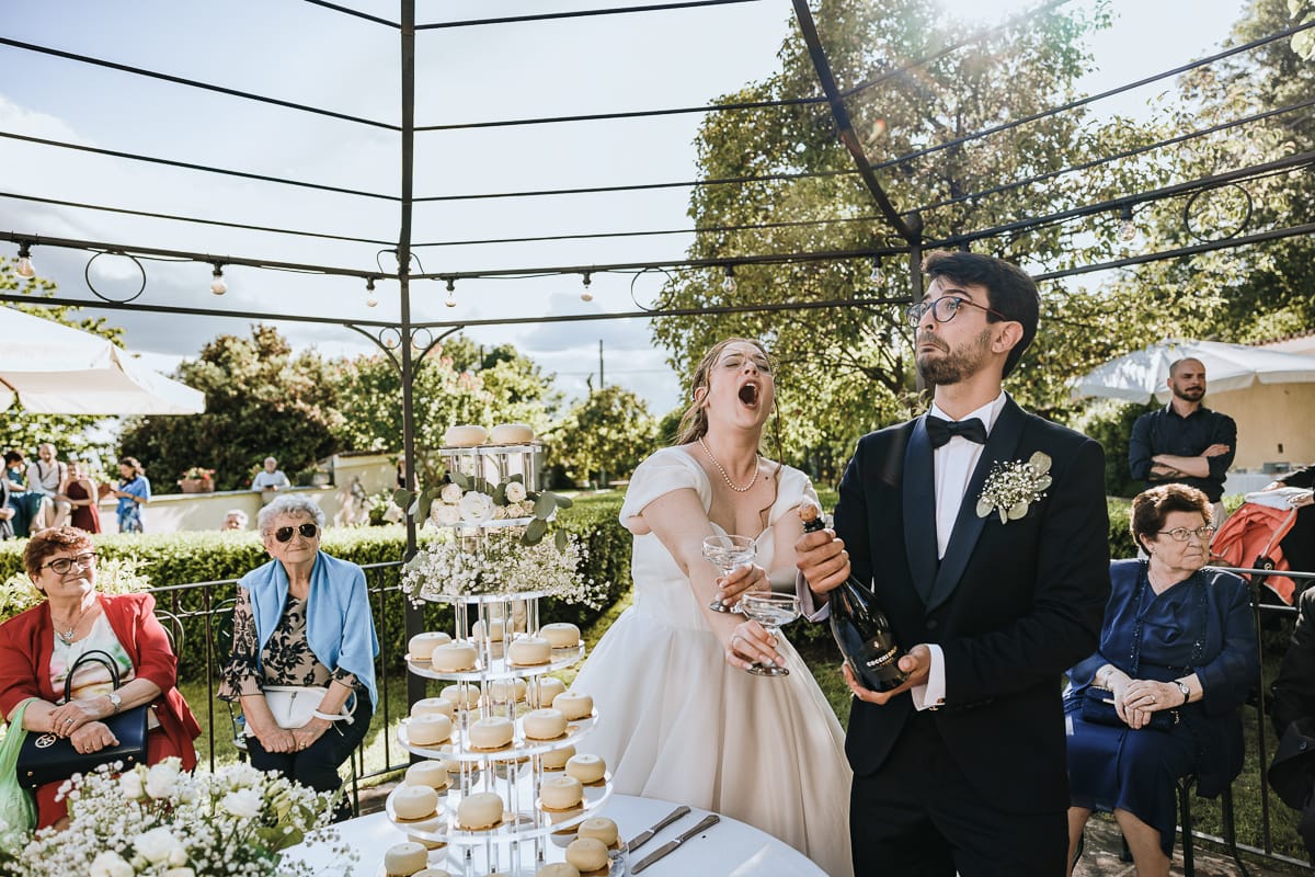 Fotografo matrimonio Torino. Taglio della torta sotto un gazebo nel giardino di Villa Ala. Apertura della bottiglia. gli sposi hanno un'espressione buffa.
