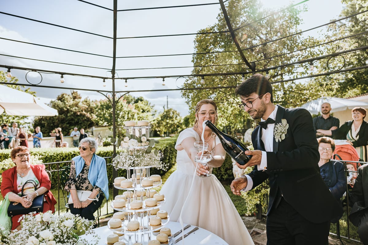 Fotografo matrimonio Torino. Taglio della torta sotto un gazebo nel giardino di Villa Ala. Lo sposo vera lo spumante nelle coppe.