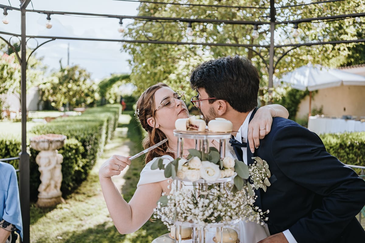Fotografo matrimonio Torino. Taglio della torta sotto un gazebo nel giardino di Villa Ala. La sposa fa finta di puntare il coltello alla gola dello sposo.