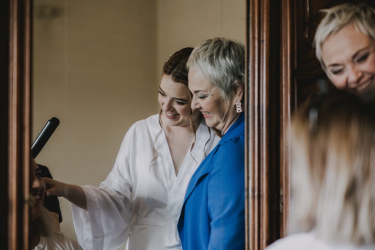 Preparazione della sposa a Madonna della Scala. Sposa e mamma riflesse in uno specchio guardano la sorella che viene pettinata.
