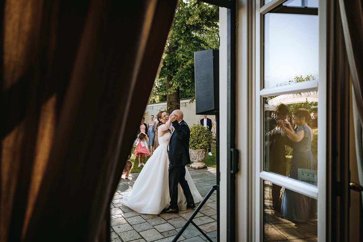 Fotografo matrimonio Torino. Ballo degli sposi con i genitori nel patio di Villa Ala. sposa con il padre e sposo con la madre riflessi in una porta finestra.