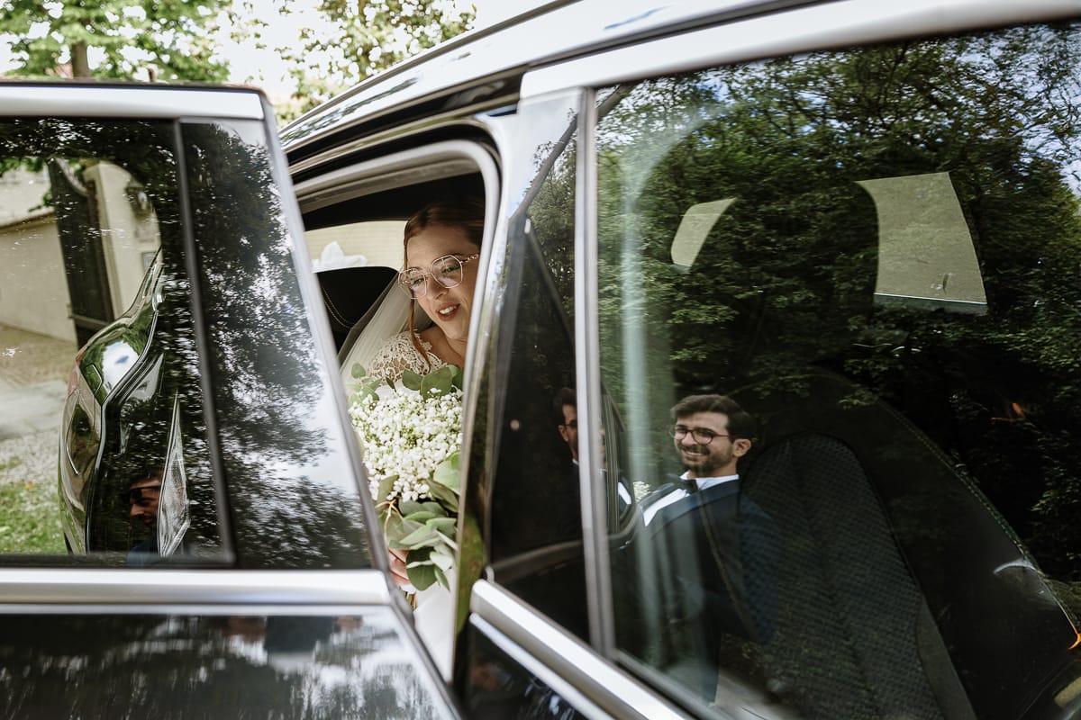 Matrimonio a Villa Ala di Rivalba (TO) Sposa scende dall'auto. Sposo fuori riflesso nel vetro del finestrino.