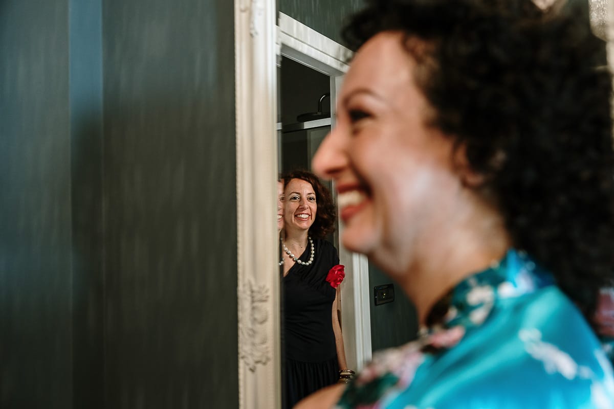 Fotografo matrimonio in Monferrato. Matrimonio di Serena e Fabrizio. Preparazione a Baldissero Torinese. Sorella della sposa esce da una stanza e guarda la sposa sfocata in primo piano.