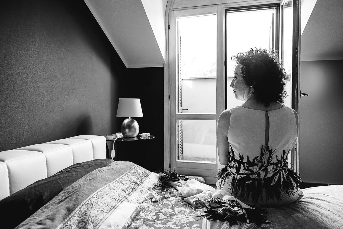 Fotografo matrimonio in Monferrato. Matrimonio di Serena e Fabrizio. Preparazione a Baldissero Torinese. Sposa seduta sul letto di schiena. bianco e nero.