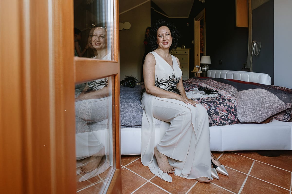 Fotografo matrimonio in Monferrato. Matrimonio di Serena e Fabrizio. Preparazione a Baldissero Torinese. Sposa seduta sul letto riflessa nella porta finestra.