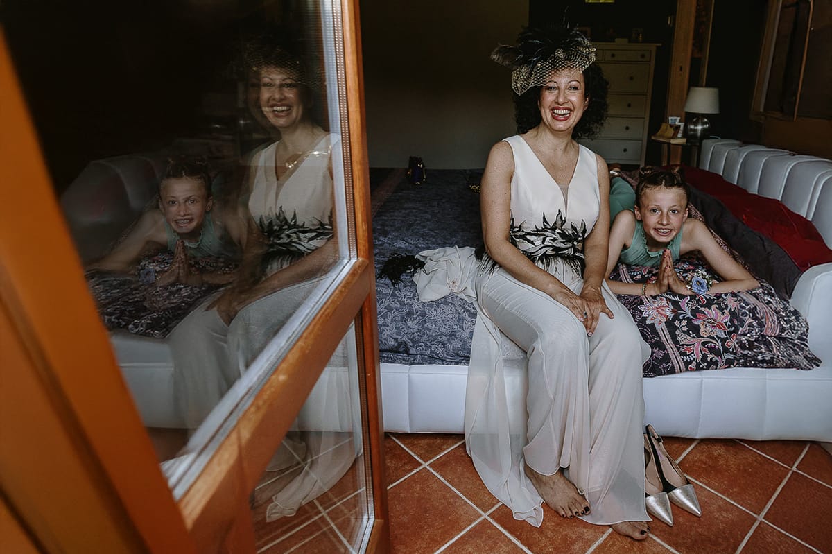 Fotografo matrimonio in Monferrato. Matrimonio di Serena e Fabrizio. Preparazione a Baldissero Torinese. Sposa seduta sul letto con la nipotina. entrambe riflesse nel vetro della porta finestra.