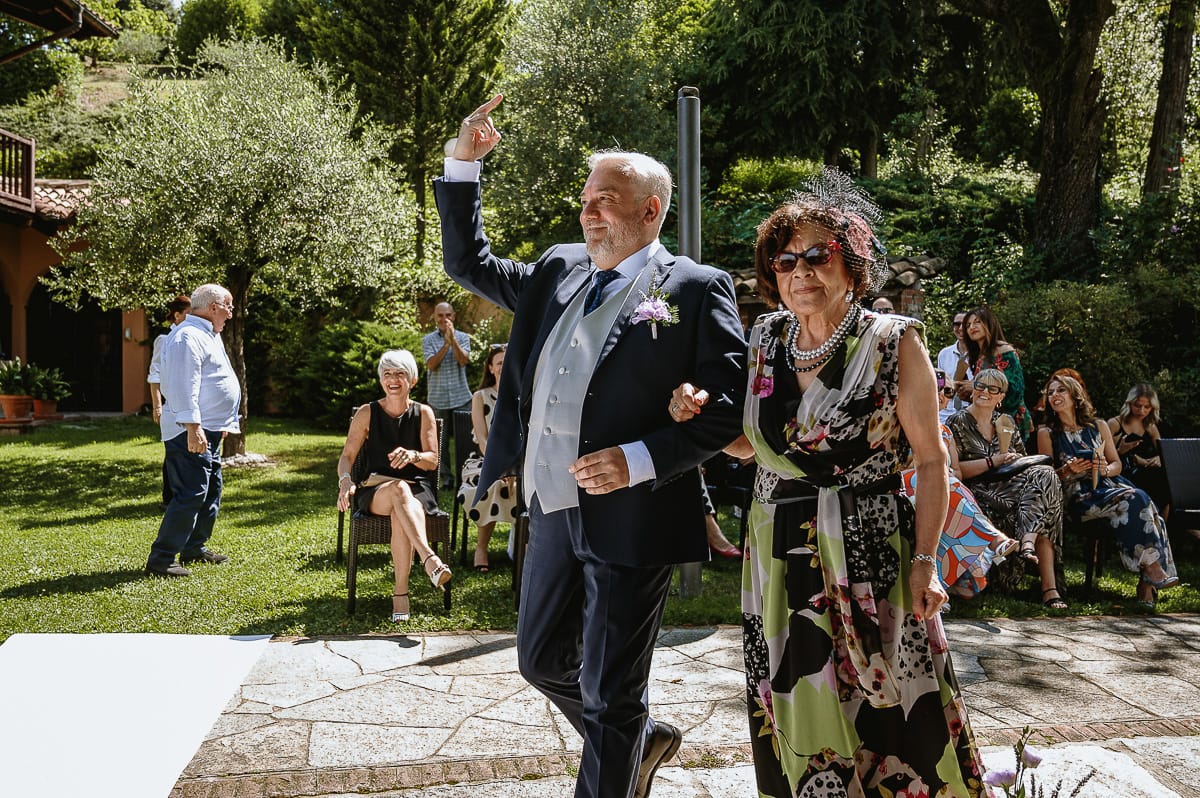 Fotografo matrimonio in Monferrato. Matrimonio di Serena e Fabrizio al Vecchio Castagno di Cocconato d'Asti.Sposo si dirige verso la zona della cerimonia al braccio della suocera mostrando il dito medio scherzando.