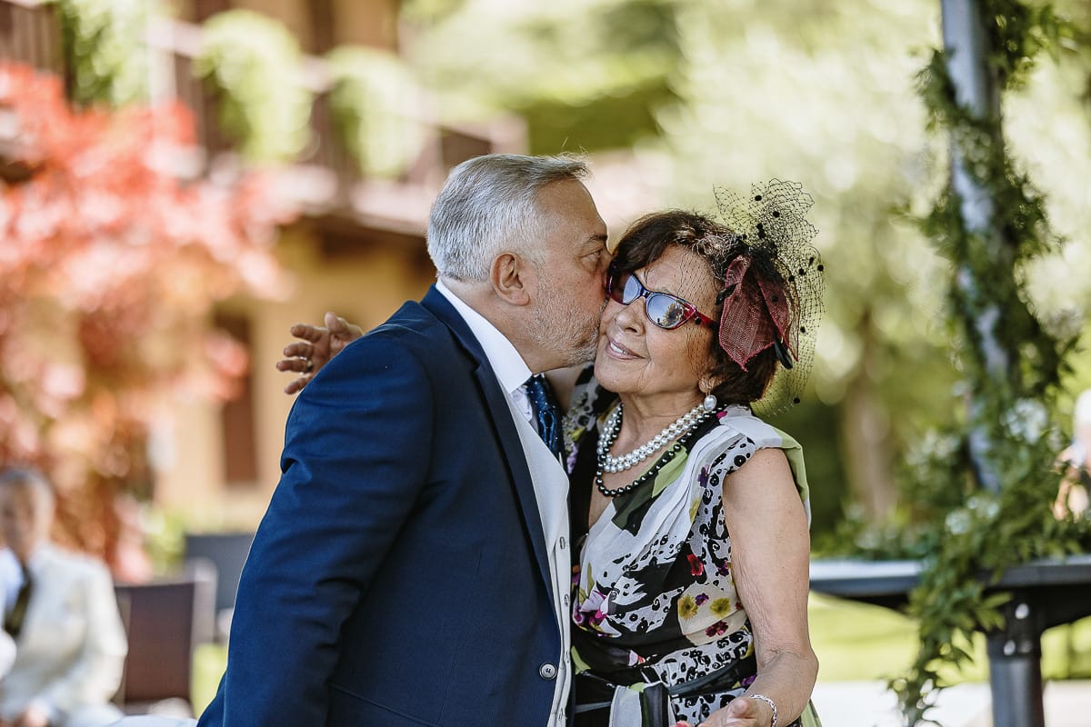 Fotografo matrimonio in Monferrato. Matrimonio di Serena e Fabrizio al Vecchio Castagno di Cocconato d'Asti.Sposo si dirige verso la zona della cerimonia al braccio della suocera. Sposo bacia la suocera.