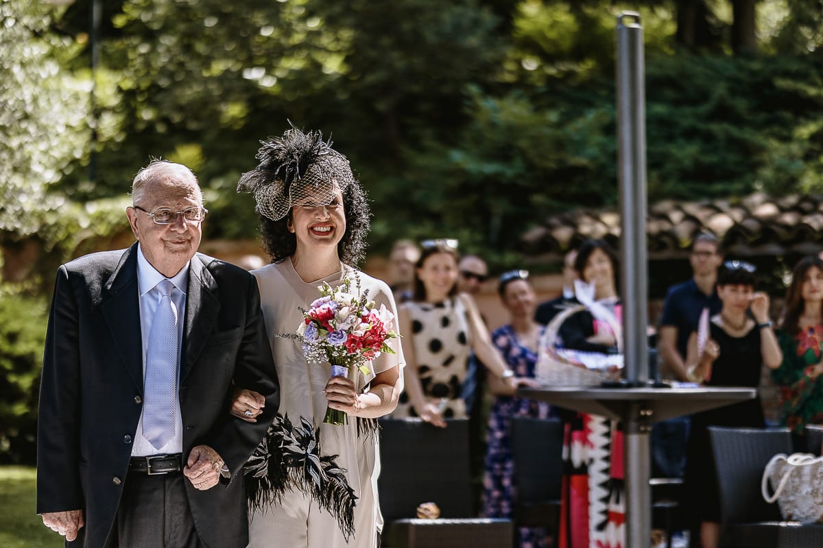 Fotografo matrimonio in Monferrato. Matrimonio di Serena e Fabrizio al Vecchio Castagno di Cocconato d'Asti. Sposa si dirige verso il luogo della cerimonia al braccio del padre.