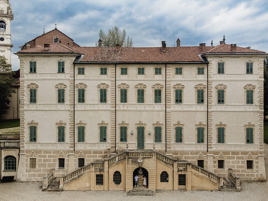 Pre-wedding at Castello di Santena. Fabiola and Cristian under the castle's staircase, in a niche near a statue, look at each other. Panoramic photo of the castle taken by drone.  