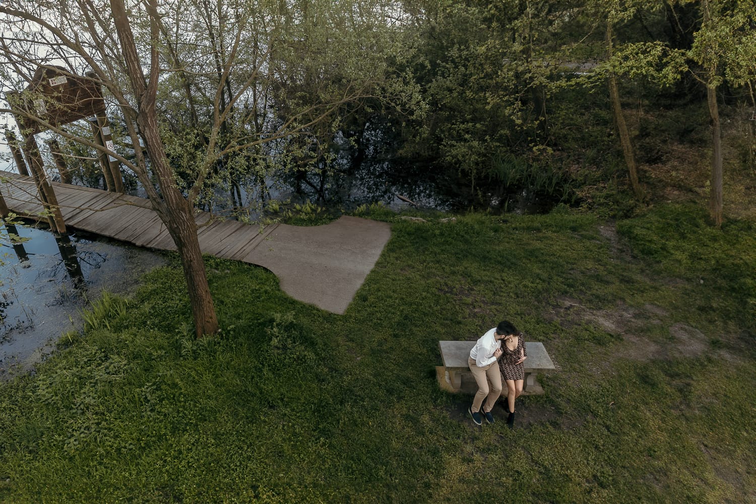 Pre-wedding al Lago di Viverone di Ilaria e Luca. Foto aerea dal drone della coppia seduta su di una panchina in pietra davanti alla zona delle palafitte.