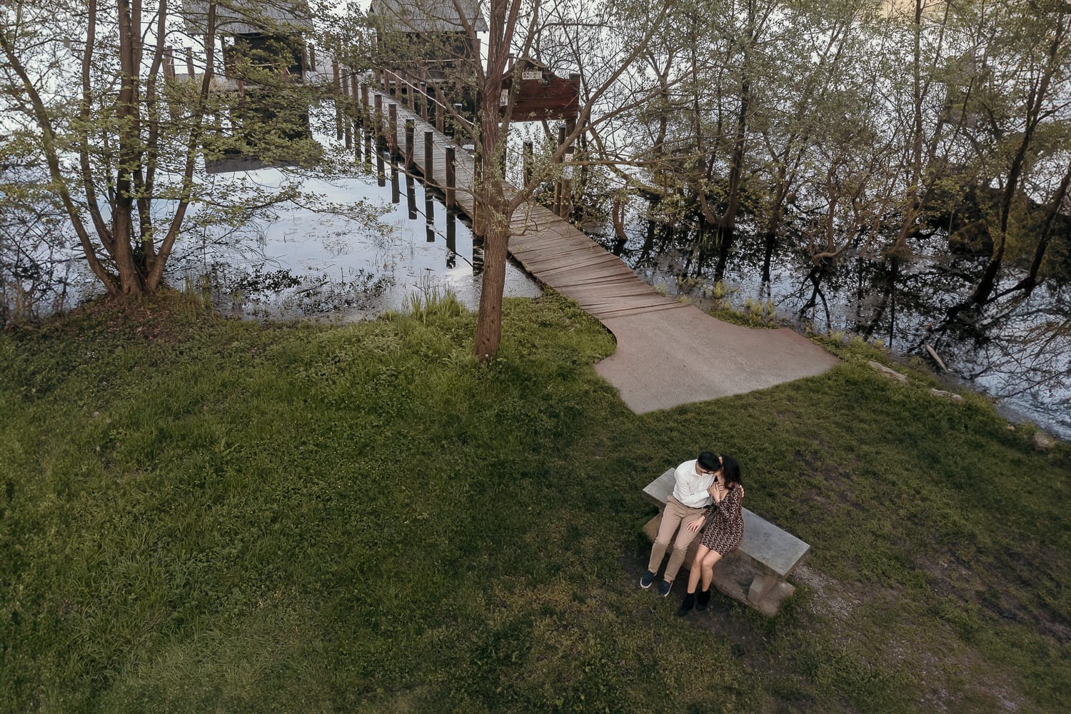 Pre-wedding al Lago di Viverone di Ilaria e Luca. Foto aerea dal drone della coppia seduta su di una panchina in pietra davanti alla zona delle palafitte.