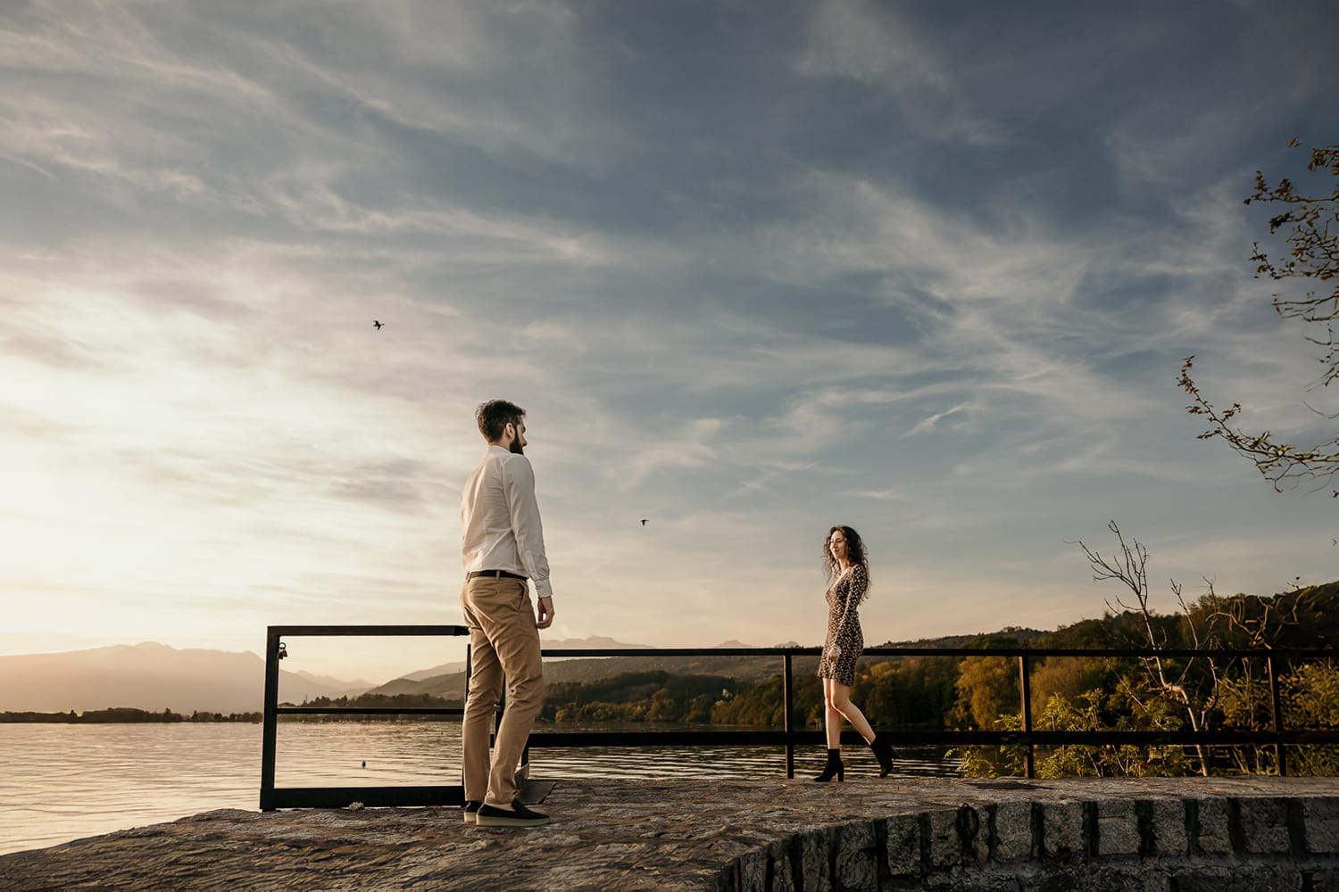 Pre-wedding al Lago di Viverone di Ilaria e Luca. Foto della coppia che cammina uno verso l'altra sul pontile di pietra sul lago al tramonto.