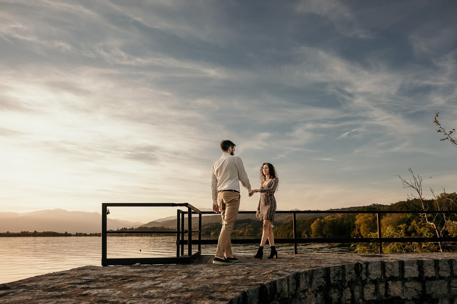 Pre-wedding al Lago di Viverone di Ilaria e Luca. Foto della coppia che cammina uno verso l'altra sul pontile di pietra sul lago al tramonto.