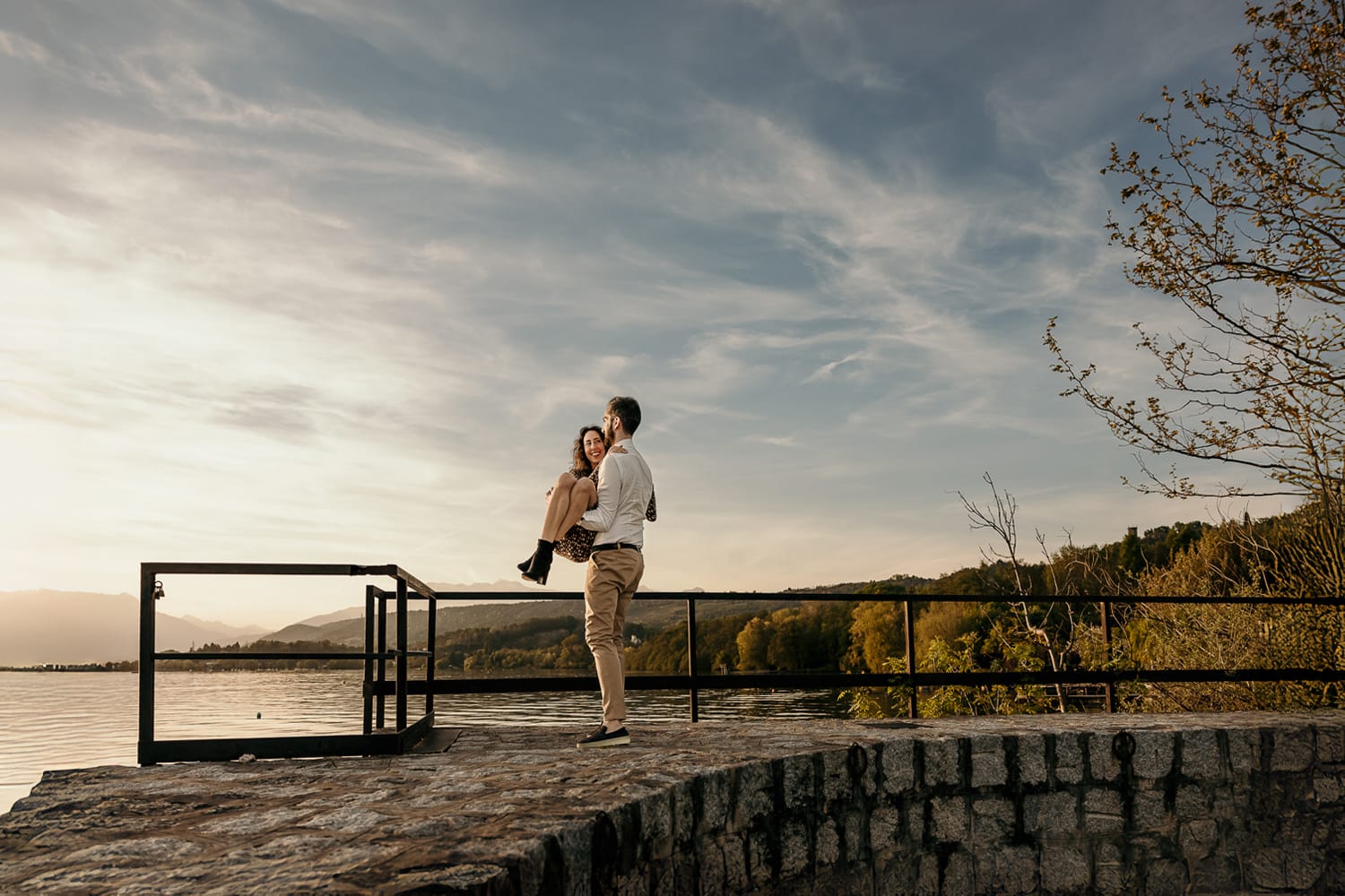 Pre-wedding al Lago di Viverone di Ilaria e Luca. Luca prende Ilaria in braccio sul pontile di pietra sul lago al tramonto.