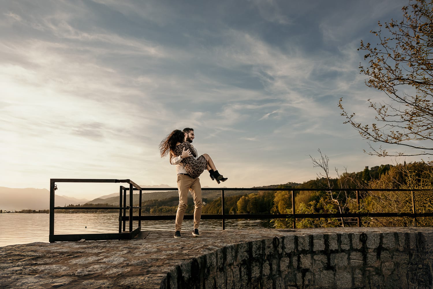 Pre-wedding al Lago di Viverone di Ilaria e Luca. Luca prende Ilaria in braccio sul pontile di pietra sul lago al tramonto.