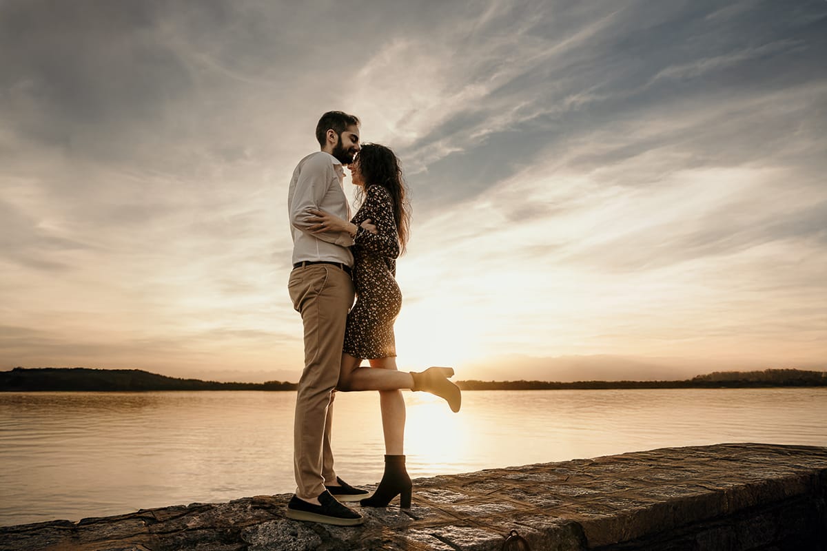 Pre-wedding al Lago di Viverone di Ilaria e Luca. Luca bacia Ilaria sulla fronte sul pontile di pietra sul lago al tramonto.