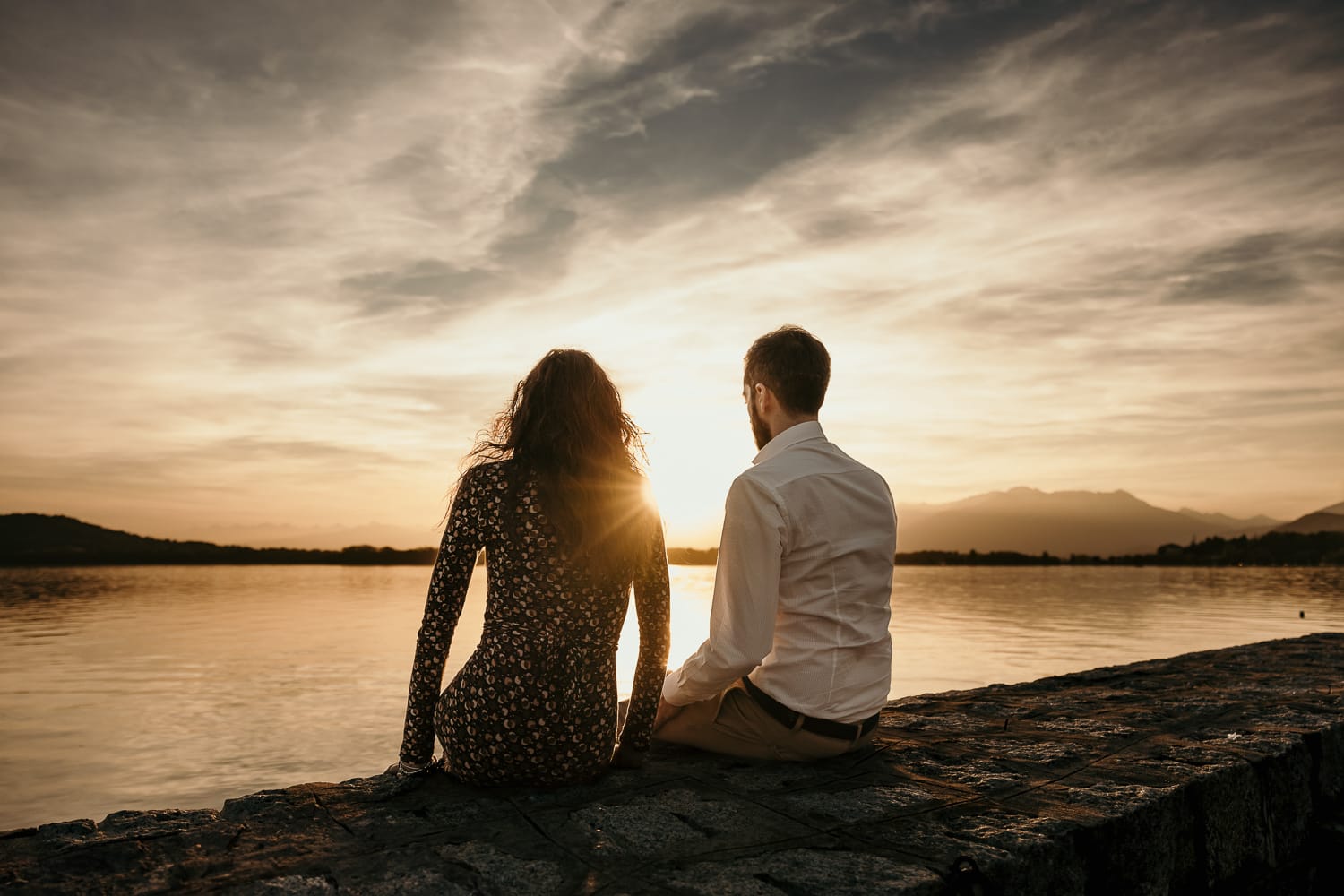 Pre-wedding al Lago di Viverone di Ilaria e Luca. Ilaria e Luca sono seduti sul pontile di pietra e guardano il bellisismo tramonto sul lago. Il sole crea un flare sulla spalla destra di Ilaria.