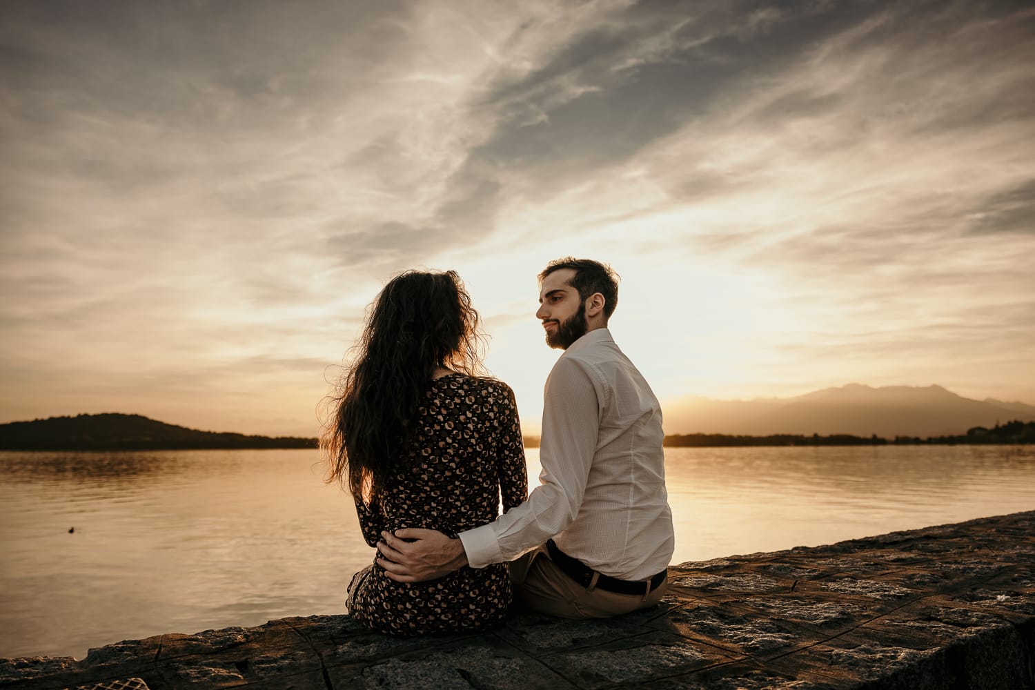 Pre-wedding al Lago di Viverone di Ilaria e Luca. La coppia si guarda mentre è seduta sul pontile di pietra davanti al bellisismo tramonto sul lago.