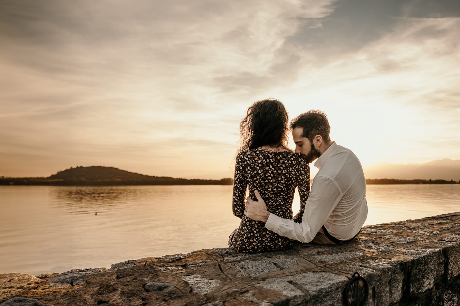 Pre-wedding al Lago di Viverone di Ilaria e Luca. La coppia è seduta sul pontile di pietra davanti al bellissimo tramonto sul lago e Luca bacia Ilaria su di una spalla.
